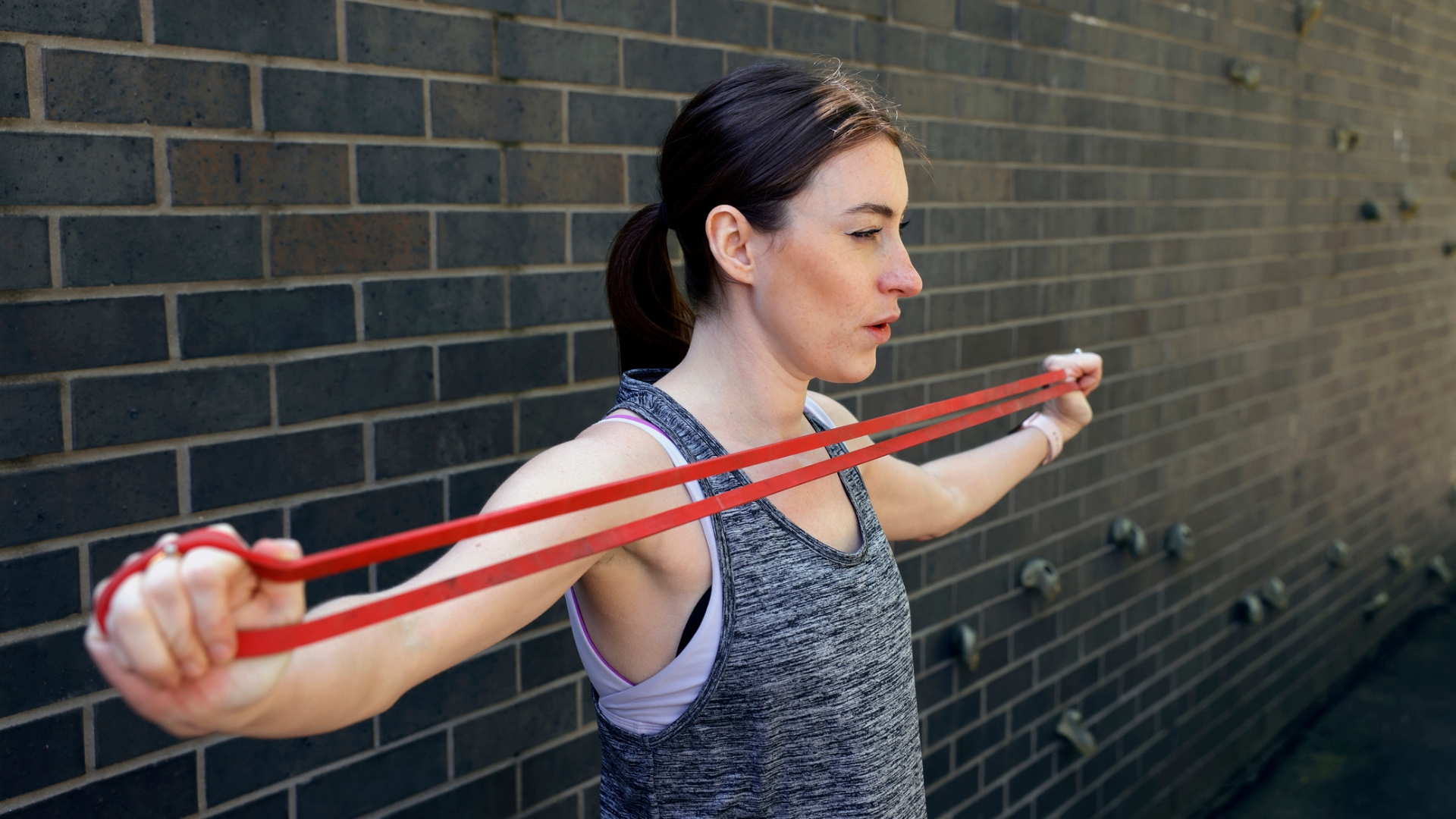 woman in a grey vest standing sideways to the camera against a brick wall stretching a red long loop resistance band across her chest
