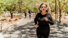 woman dressed in black sleeved top and leggings running outside with white headphones. she's on a path with trees and other people blurred in the background 