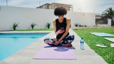 Woman sitting on yoga mat next to a pool looking at her smartphone