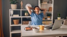 Woman sits at desk in front of open laptop, leaning back in her chair with her hands behind her head and her eyes closed