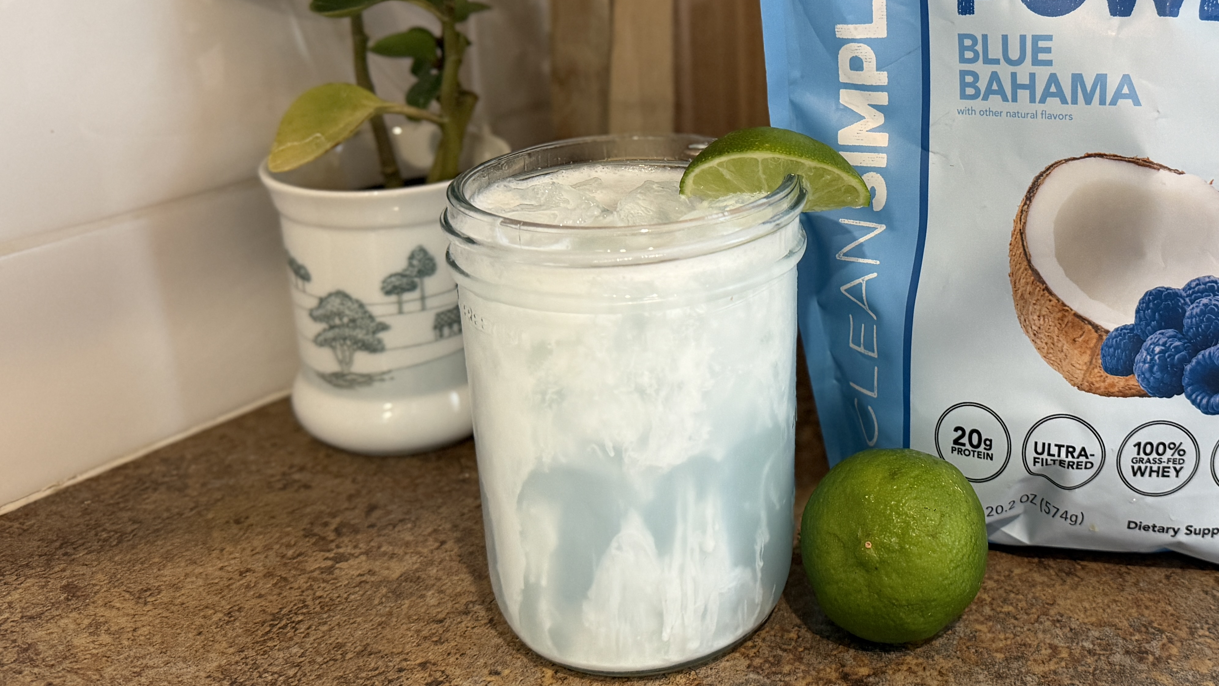 Mason jar of white liquid garnished with lime wedge. It is on a countertop next to a plant pot, whole lime, and packet of unidentified food