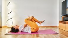 woman on wooden floor and a pink yoga mat lying on her back with her knees close to her chest in a figure-four stretch. she's wearing a white tshirt and orange leggings. 