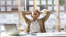 woman wearing a white shirt and beige jacket sitting at a desk with a laptop in front, her hands are behind her head mid-stretch. there's a window behind her and papers on the desk. 