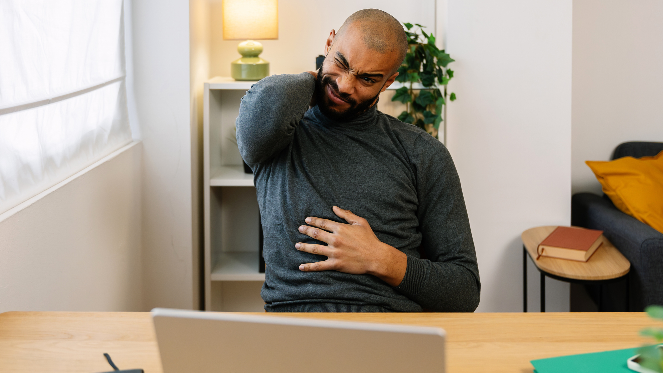 Man sitting in front of a laptop on a desk winces while he holds his neck with one hand
