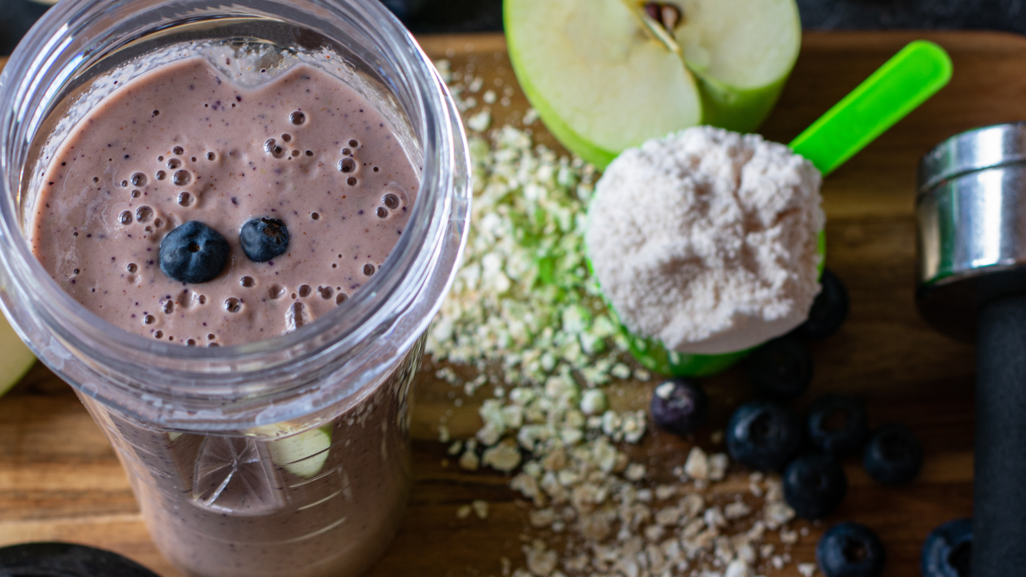 A blender cup containing a meal replacement shake with a scoop of powder, an apple and some oats on a worktop