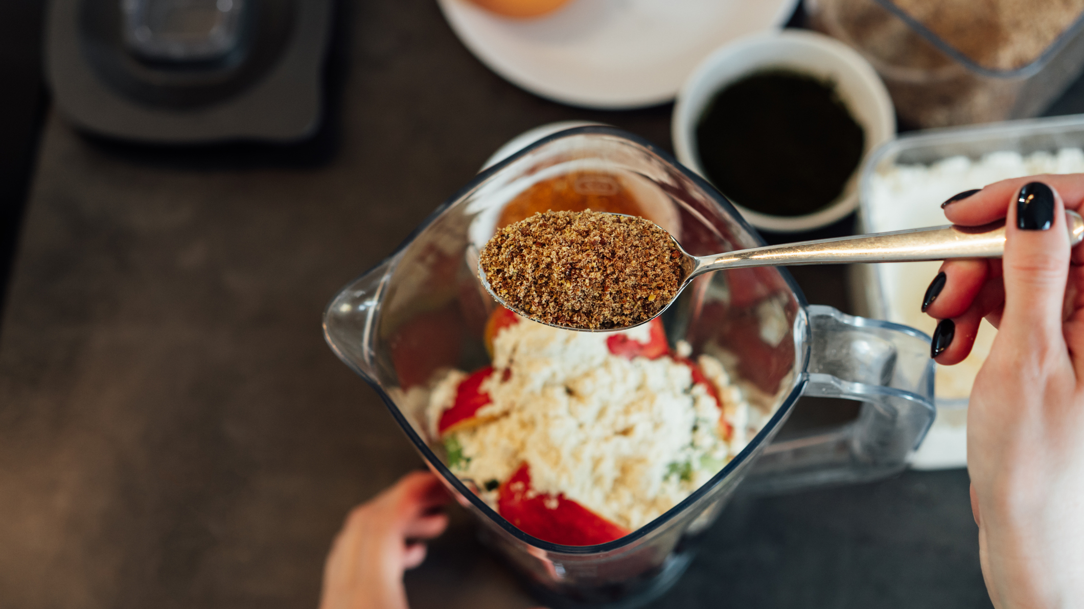 Spoon of brown powder seen above blender jug with unidentifiable ingredients in it