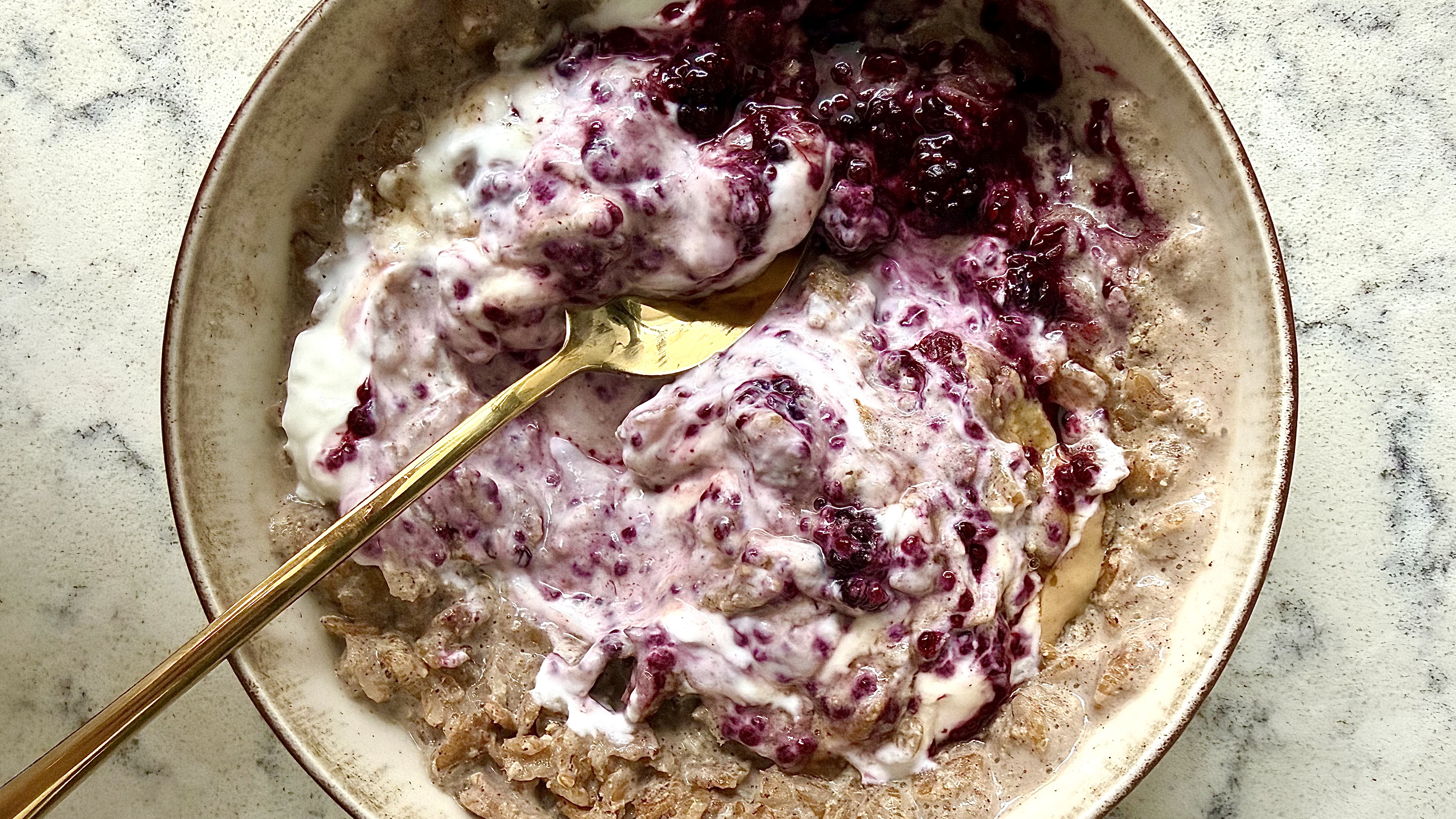 A bowl of porridge with yoghurt and jam swirled in, a brass-colored, shiny spoon rests in the bowl